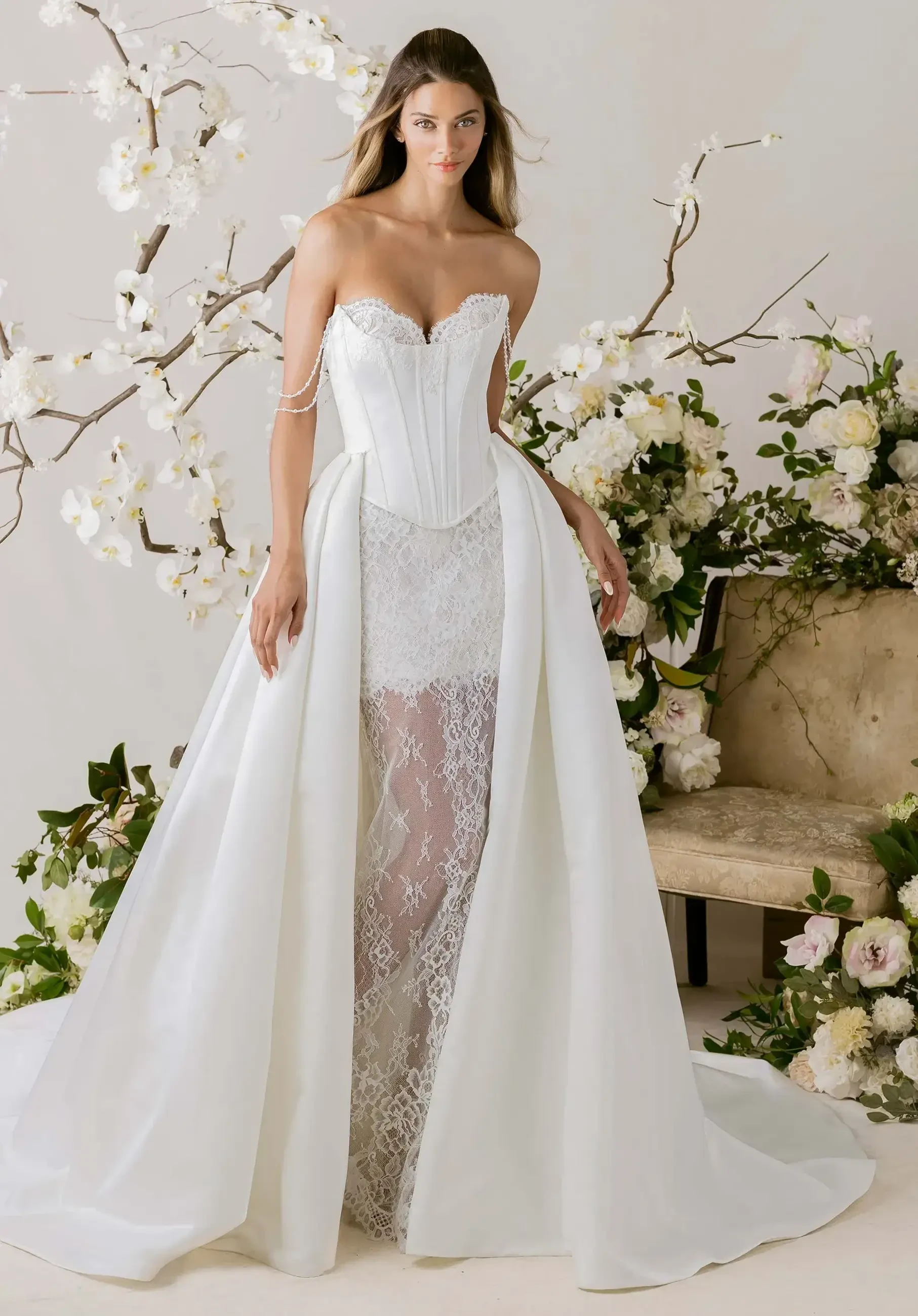 A model is wearing an elegant white wedding dress with a corset top and a flowing skirt. The dress has intricate lace details and is complemented by floral decorations in the background.
