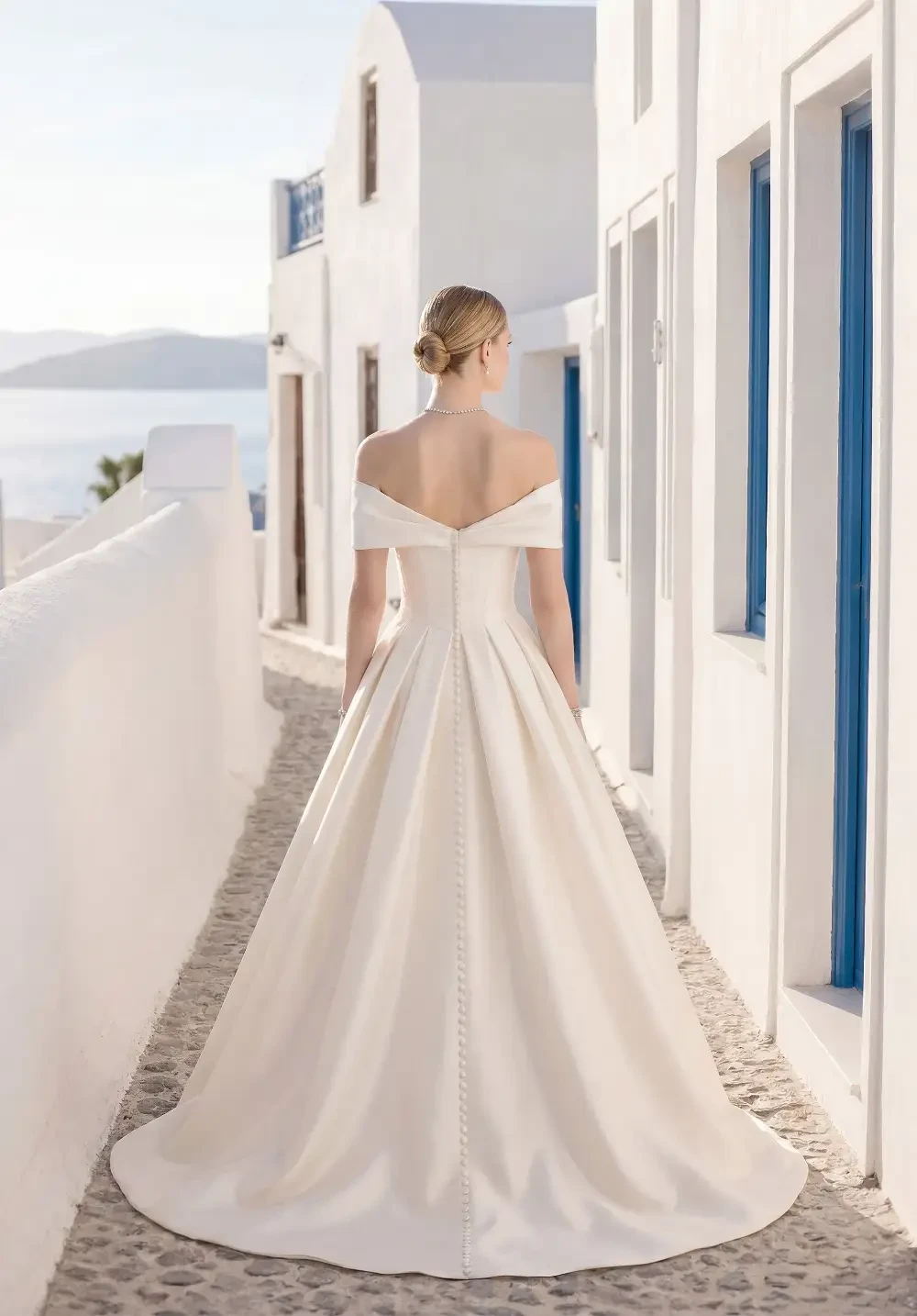 A bride in a white wedding dress standing on a cobblestone path, facing away, with white buildings and blue doors in the background.