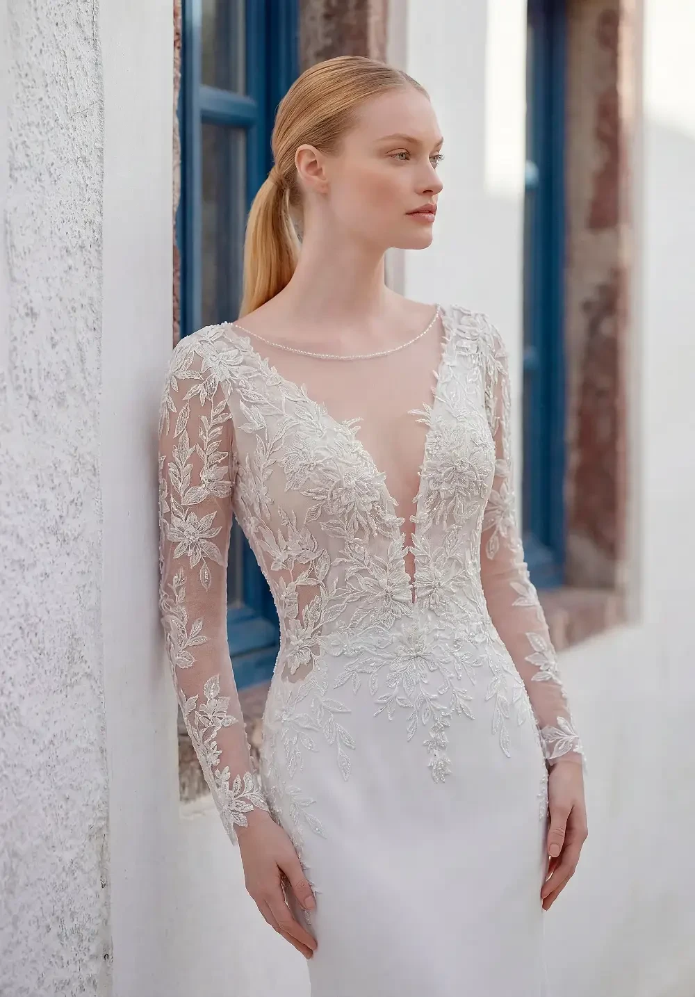 A woman in a white wedding dress with long lace sleeves and intricate embroidery, posing against a textured wall with blue window frames.