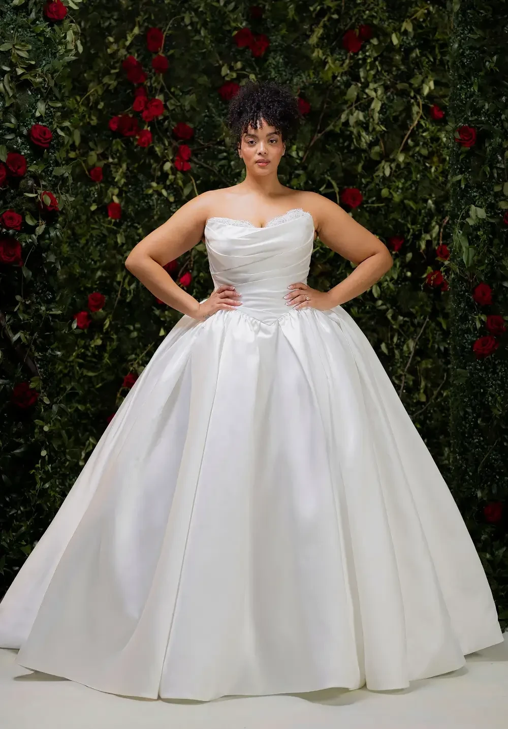 A model wearing a strapless, white ball gown stands confidently with her hands on her hips, against a backdrop of greenery and red roses.