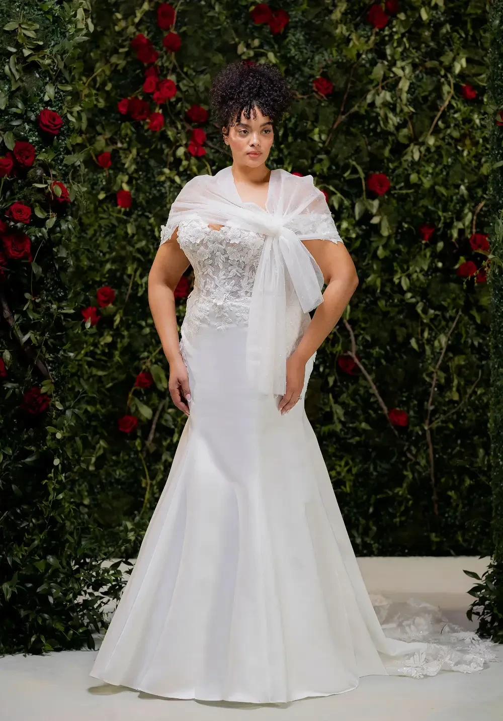 A woman wearing a white wedding dress with a lace bodice and a flowy shawl, standing in front of a backdrop of roses.