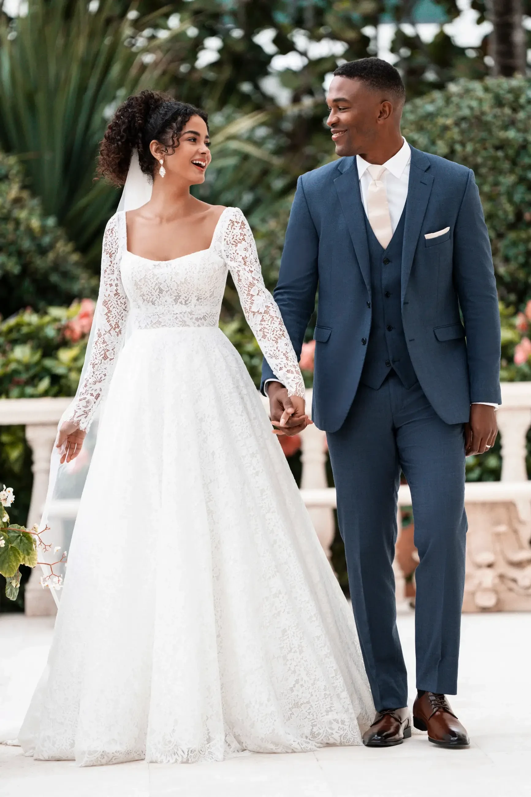 A bride in a white lace gown and veil and a groom in a blue suit with a white tie joyfully hold hands, walking outdoors amidst lush greenery.