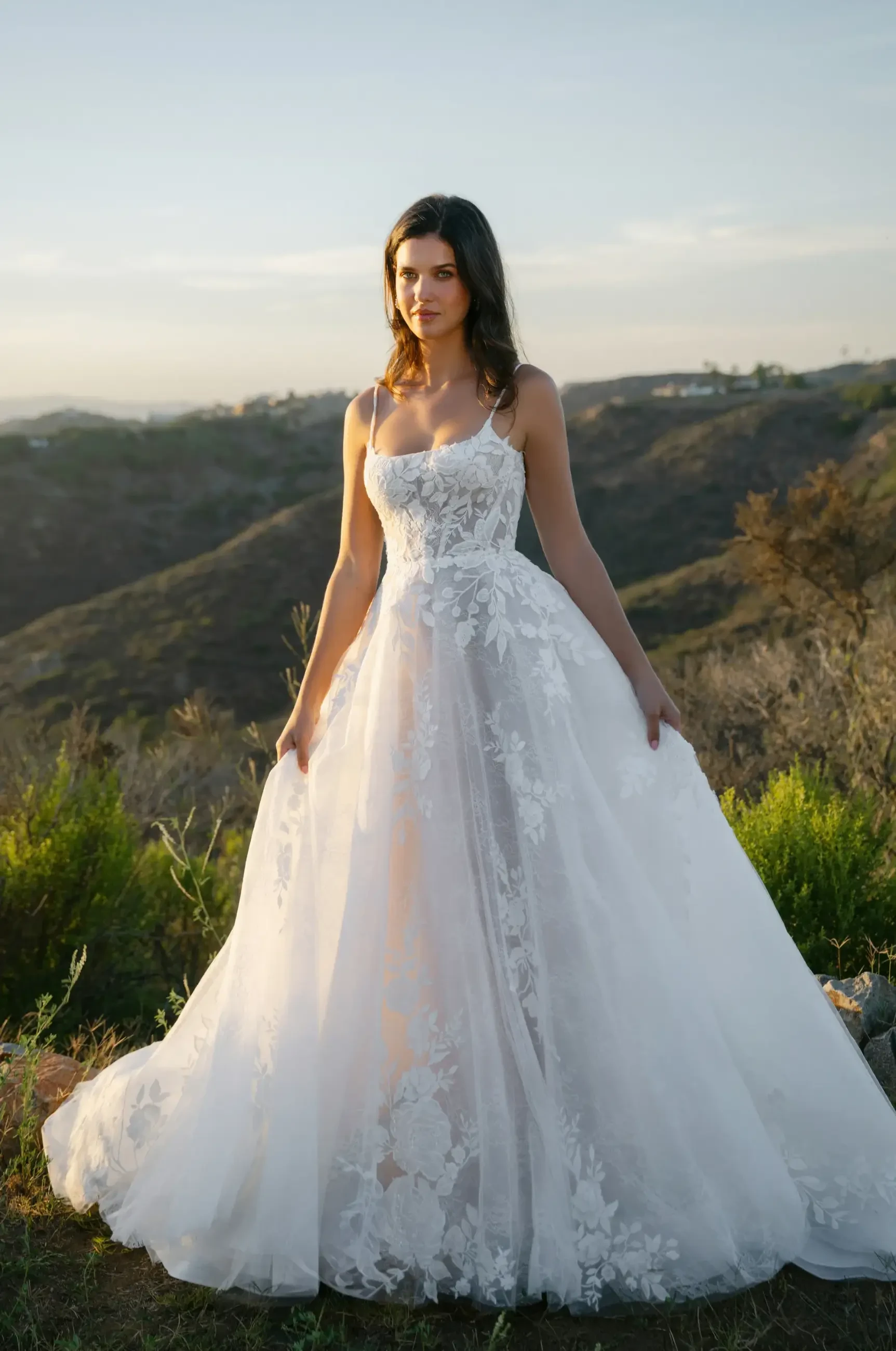 A woman stands on a hilltop wearing a flowing white lace wedding gown. The background features rolling hills and a soft, hazy sky at sunset.