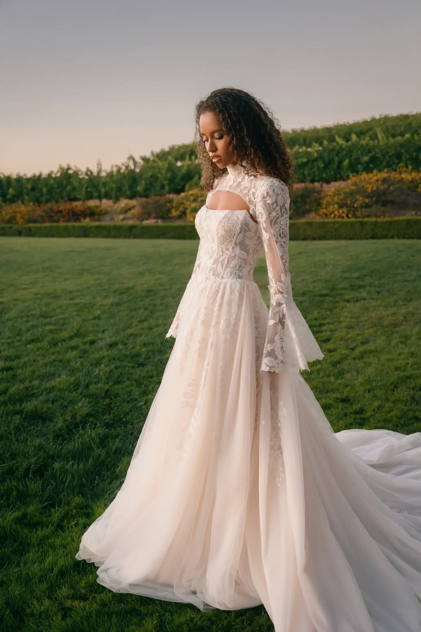 A woman in a flowing lace wedding gown, featuring sheer sleeves, stands thoughtfully on a lush green lawn at sunset, with a serene backdrop of hedges.