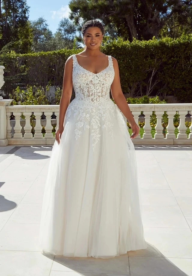A model wearing an elegant white wedding gown with off-the-shoulder sleeves and a thigh-high slit, standing in front of a floral backdrop.