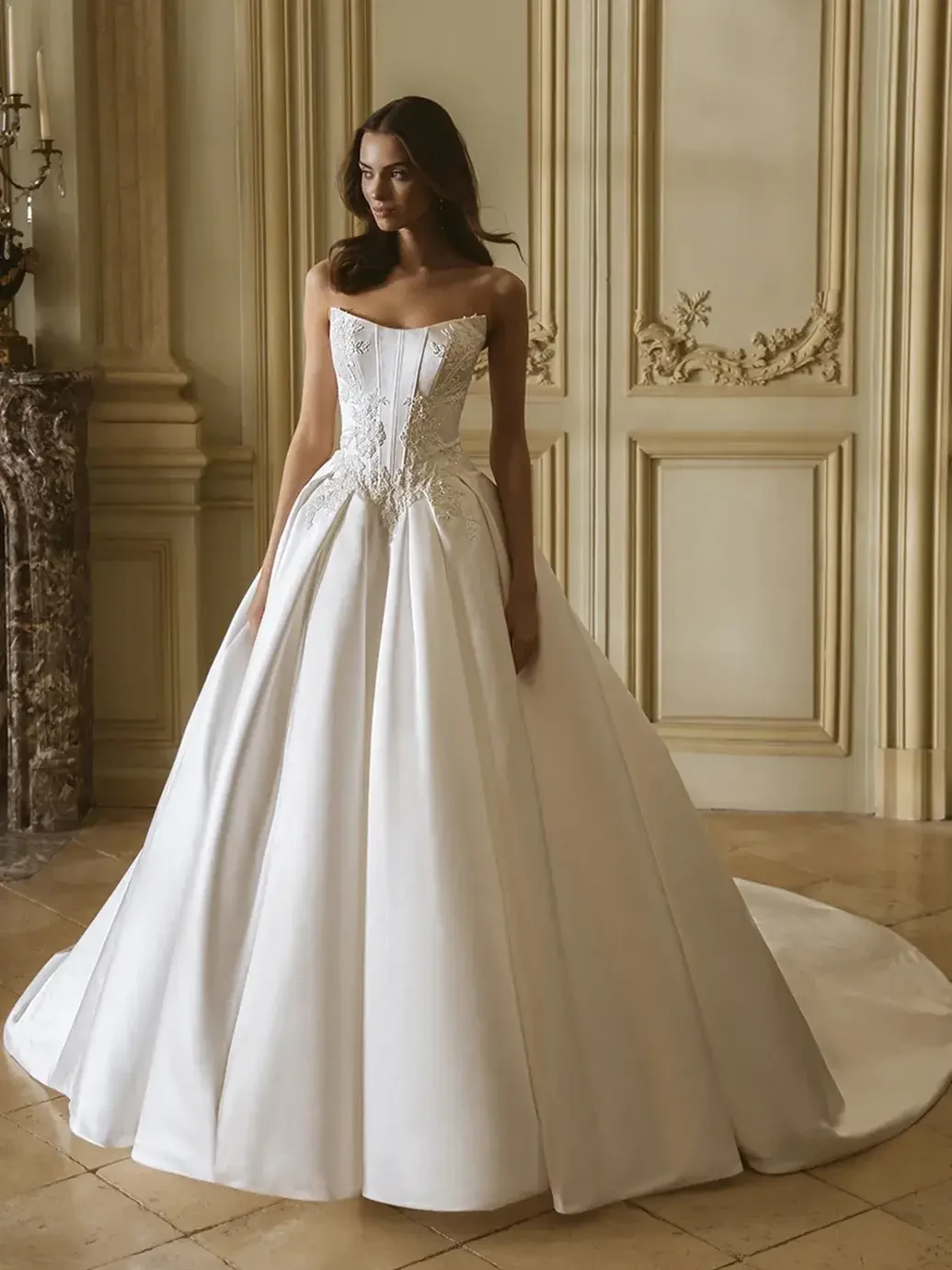 A woman wearing a strapless, wedding gown with a full skirt and intricate detailing, standing in an elegant room with ornate decor.