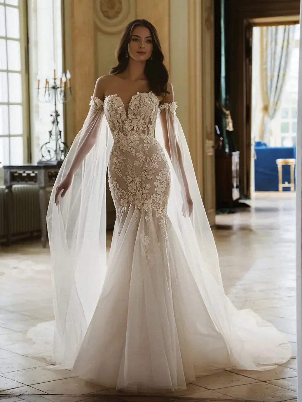 A woman in a stunning bridal gown with intricate lace details and flowing sheer sleeves, standing elegantly in a beautifully decorated room.