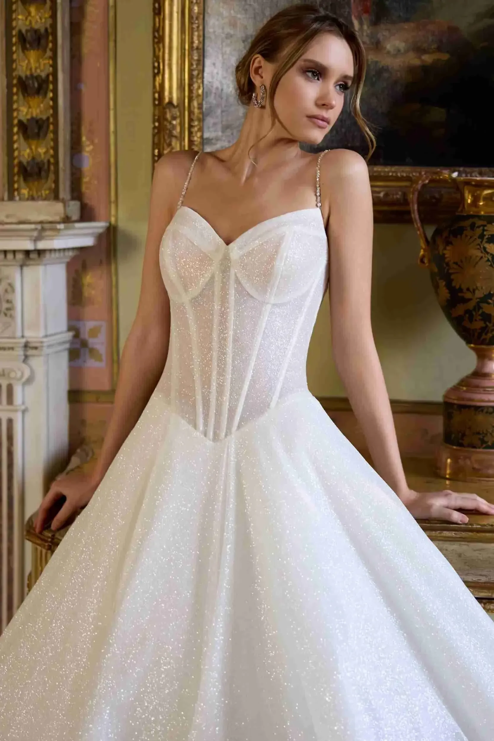 A woman in an elegant, sparkling white gown with thin straps leans on a table in a richly decorated room, exuding a serene and thoughtful mood.
