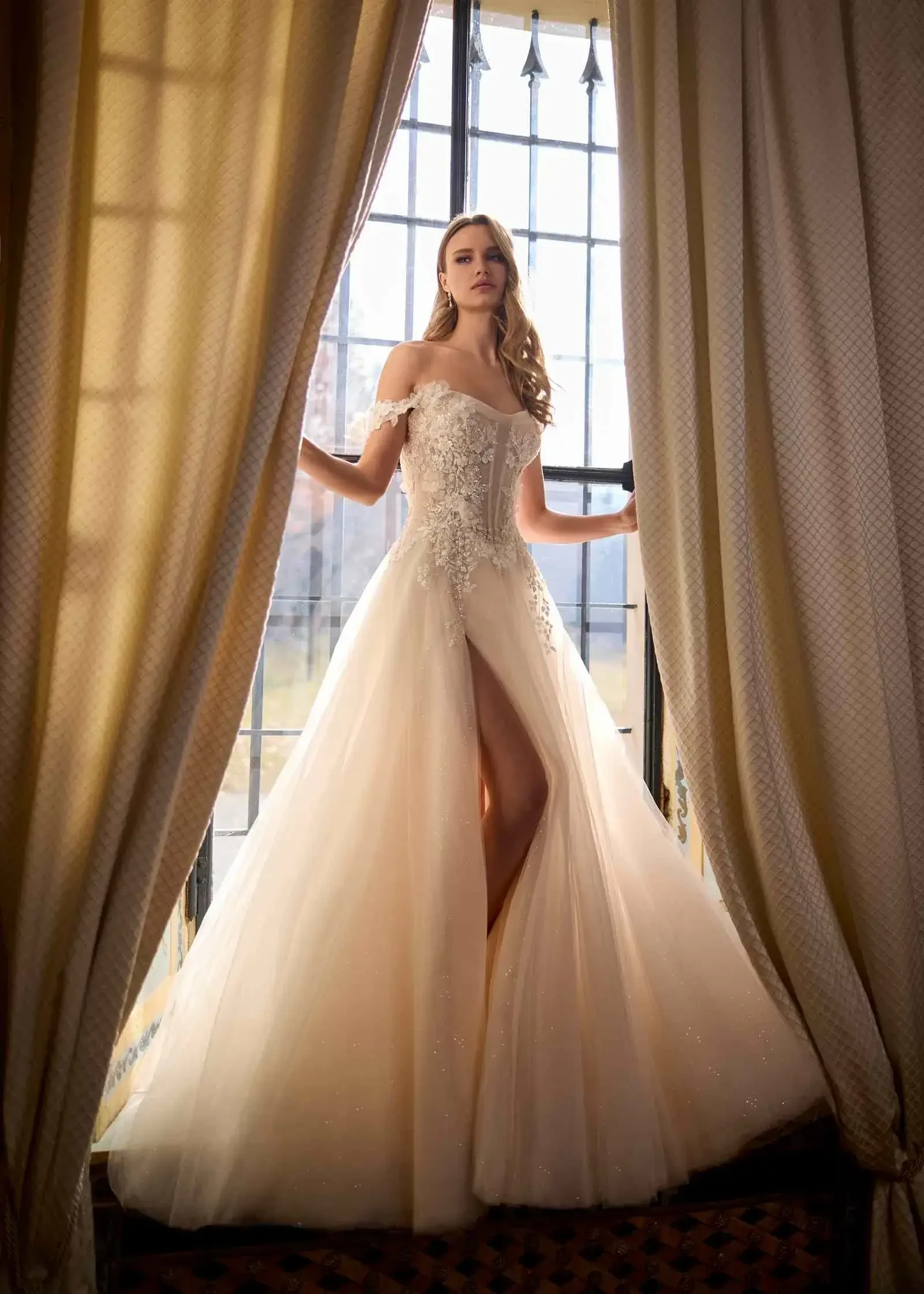 A woman stands gracefully in front of tall, sunlit windows. She is wearing an elegant, off-the-shoulder gown with lace details and a high slit. Ivory curtains frame the scene, adding a classic, serene atmosphere.